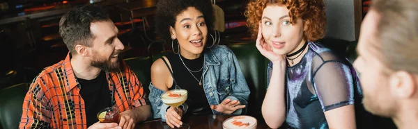 Emocionadas mujeres interracial sosteniendo cócteles cerca de hombres borrosos en el bar, pancarta - foto de stock