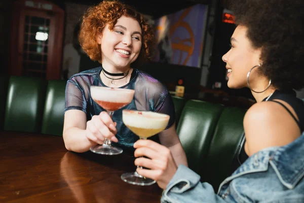 Улыбающиеся межрасовые девушки, держащие размытые коктейли в баре — стоковое фото