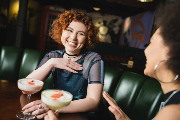 Femme rousse positive tenant un cocktail trèfle club près d'un ami afro-américain dans un bar — Photo de stock