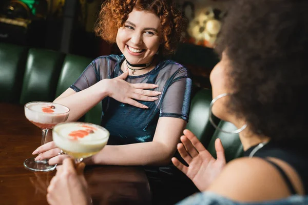 Femme insouciante tenant trèfle club cocktail près flou afro-américaine copine dans le bar — Photo de stock