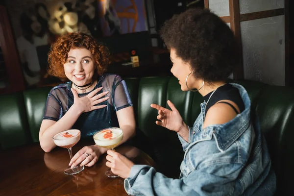 Cheerful interracial girlfriends talking near foam cocktails in bar at night — Stock Photo