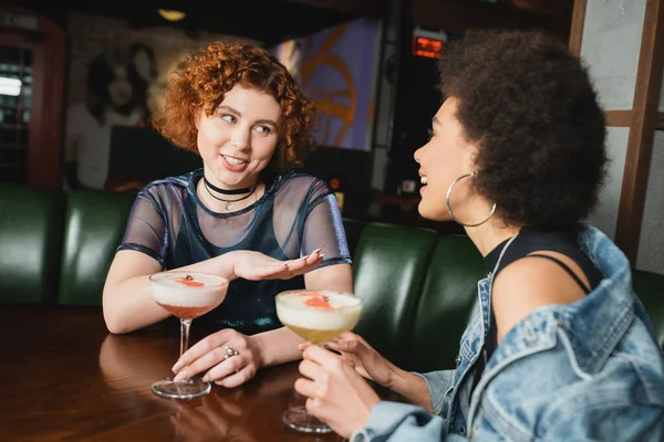 Smiling red haired woman talking to african american girlfriend near cocktails in bar — Stock Photo