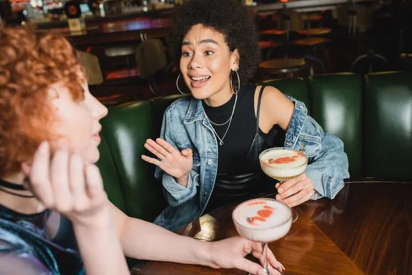 Femme afro-américaine excitée parlant à un ami flou près de cocktails en mousse dans le bar — Photo de stock