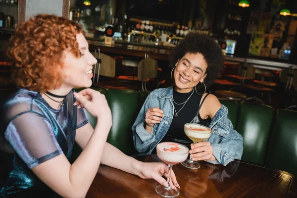 Посміхнена американка, яка дивиться на розмиту дівчину, яка розмовляє біля коктейлів у барі. — стокове фото