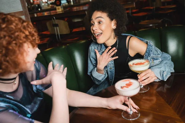Здивований афроамериканець тримає в руках кисло-рожевий коктейль і розмовляє з розмитою дівчиною в барі — стокове фото