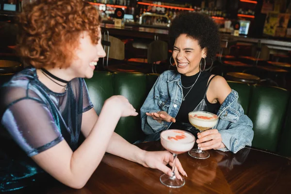 Happy multiethnic friends laughing and talking near foam cocktails in bar — Stock Photo