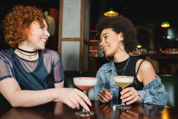 Cheerful interracial friends holding foam cocktails in bar at night — Stock Photo