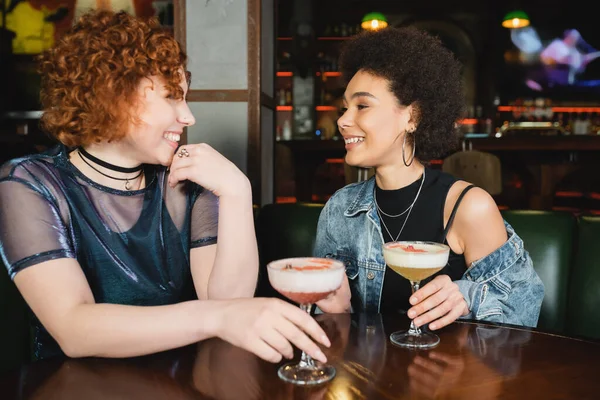 Jóvenes novias multiétnicas hablando cerca de cócteles de espuma en el bar - foto de stock