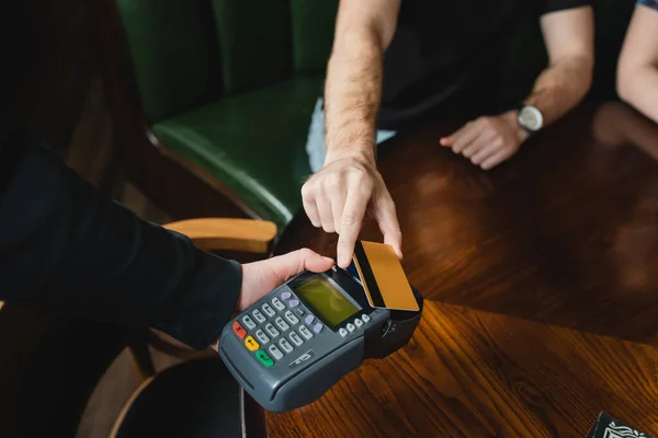 Vista cortada do garçom segurando terminal de pagamento perto do homem com cartão de crédito no bar — Fotografia de Stock