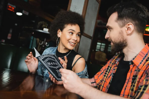 Lächelnde Afroamerikanerin im Gespräch mit einem bärtigen Freund mit Menü in der Bar — Stockfoto