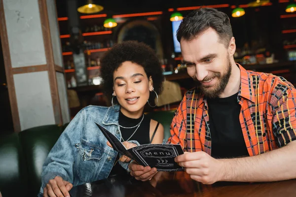 Amigos multiétnicos alegres em roupas casuais olhando para o menu no bar à noite — Fotografia de Stock