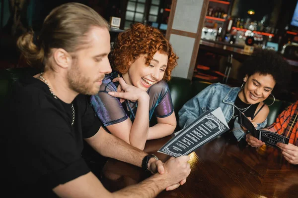 Femme rousse positive assise près d'amis interraciaux regardant le menu dans le bar — Photo de stock