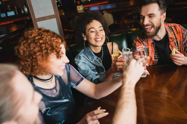 Amigos interracial positivos sosteniendo vasos de tequila y lima fresca en el bar - foto de stock