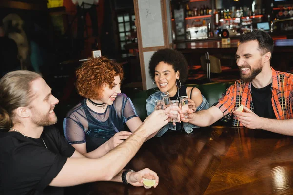 Excited multiethnic friends holding fresh lime and clinking tequila in bar — Stock Photo