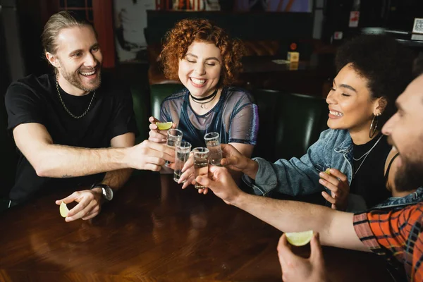 Amigos multiétnicos despreocupados tintineando tequila y sosteniendo lima en el bar por la noche - foto de stock