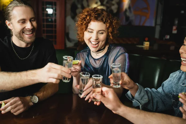 Allegro amici multietnici in possesso di shot di tequila e lime vicino rossa donna nel bar — Foto stock