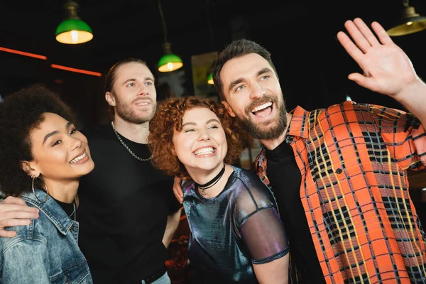 Smiling man waving hand and hugging happy multiethnic friends while waving hand in bar — Stock Photo