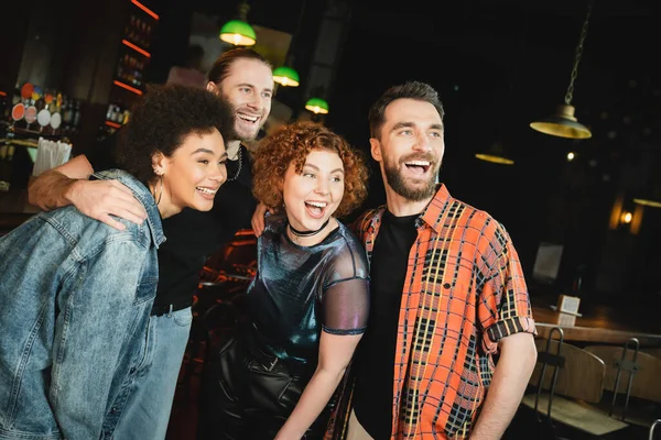 Cheerful interracial friends hugging and looking away in bar at night — Stock Photo