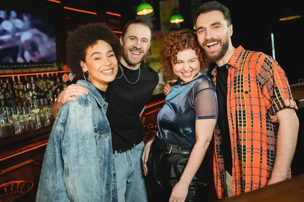 Positive bearded men hugging smiling interracial friends in bar at night — Stock Photo