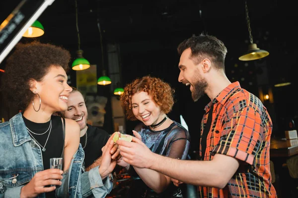 Amigos alegres sosteniendo lima y tequila cerca de un amigo afroamericano en el bar - foto de stock