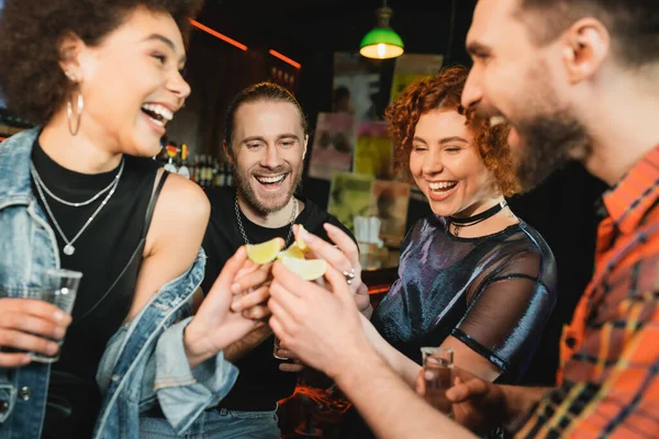 Excited interracial friends holding sour lime and tequila shots in bar — Stock Photo