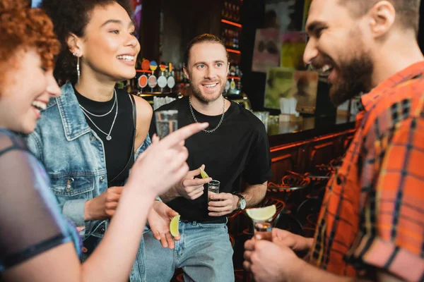 Cheerful interracial friends with tequila shots talking in bar at night — Stock Photo