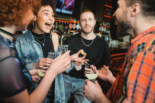 Femme afro-américaine excitée tenant un tir de tequila près d'amis flous dans un bar — Photo de stock
