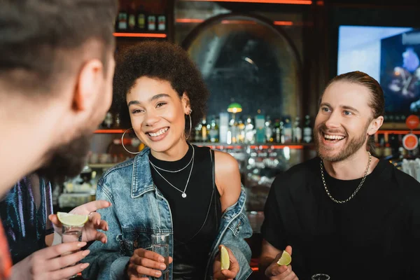 Alegre mujer afroamericana sosteniendo lima y tequila cerca de amigos en el bar - foto de stock