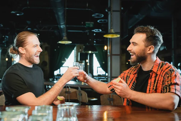 Весёлые друзья-мужчины держат лайм и звенят текилой в баре — стоковое фото