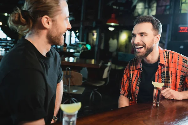 Positive bearded men talking near shots of tequila with lime in bar at night — Stock Photo