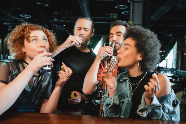 Amigos multiétnicos tomando tragos de tequila y sosteniendo lima fresca en el bar - foto de stock