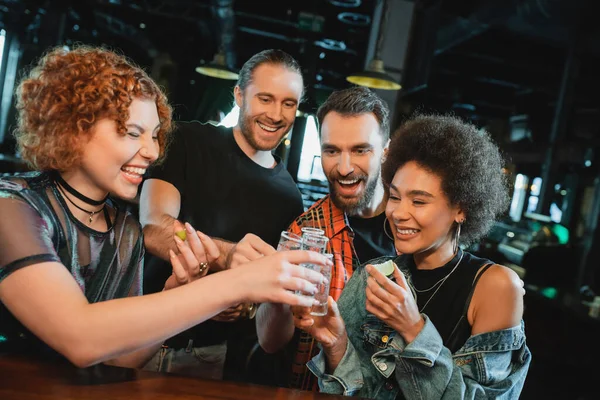 Smiling multiethnic friends toasting with tequila shots with salt and lime in bar — Stock Photo