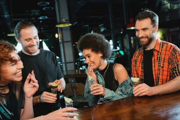 Cheerful african american woman holding glass of tequila with lime near friends in bar — Stock Photo