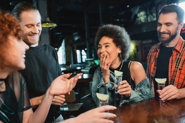 Mujer afroamericana riendo cerca de amigos con tragos de tequila en bar - foto de stock