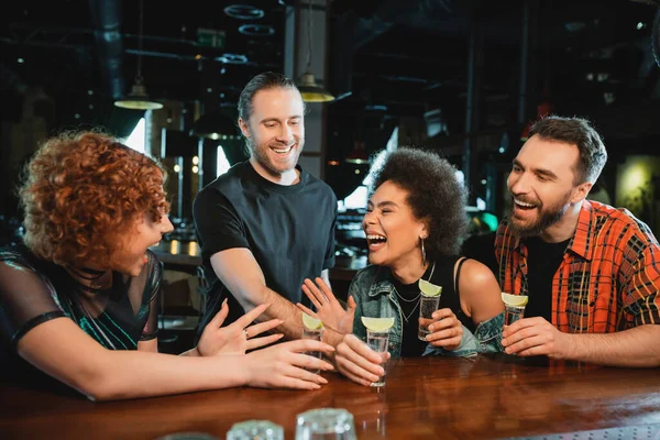 Cheerful interracial friends with tequila talking and laughing near stand in bar — Stock Photo