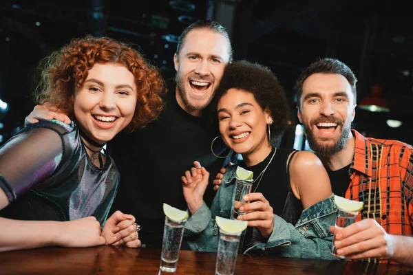 Positivos amigos interracial sosteniendo tomas de tequila y mirando a la cámara en el bar - foto de stock