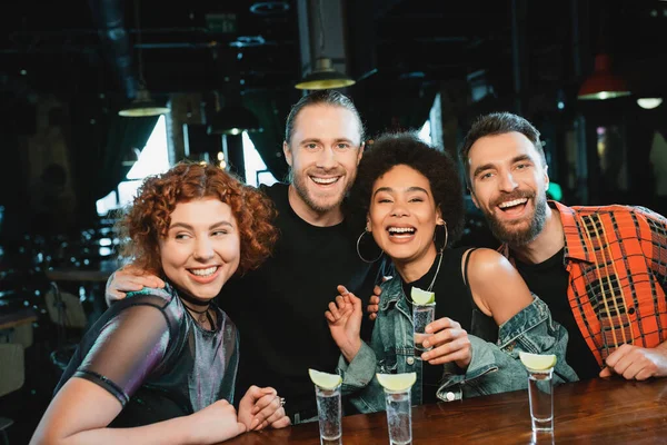 Cheerful interracial friends looking at camera near glasses with tequila in bar — Stock Photo