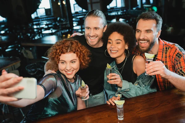 Cheerful interracial friends with tequila shots taking selfie on smartphone in bar — Stock Photo
