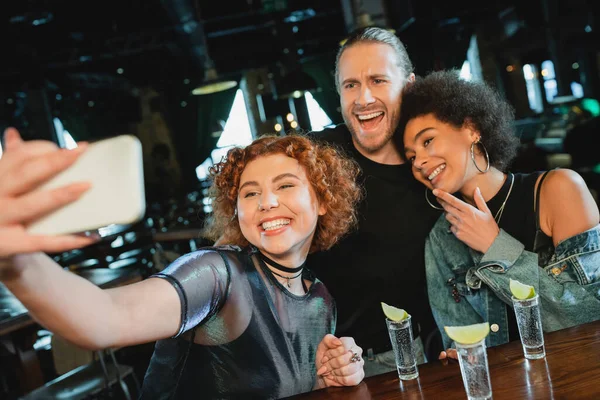 Mujer positiva tomando selfie con amigos multiétnicos cerca de tequila y lima en el bar - foto de stock