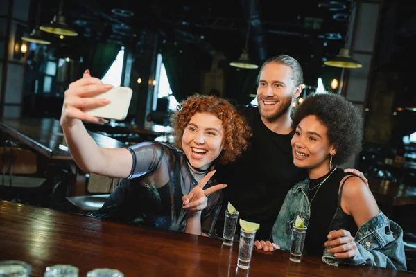 Cheerful multiethnic friends taking selfie on smartphone near tequila in bar — Stock Photo