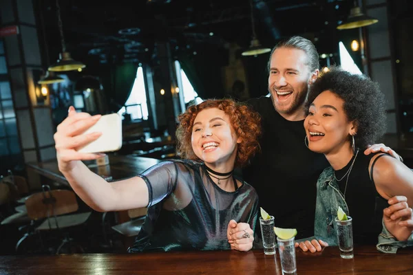Donna rossa che prende selfie con amici sorridenti vicino alla tequila nel bar — Foto stock