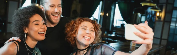 Щаслива руда жінка бере селфі на смартфон біля багатоетнічних друзів в барі, банер — стокове фото