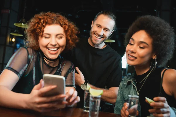 Cheerful multiethnic friends using smartphone near tequila shots in bar — Stock Photo