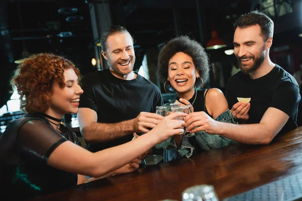 Carefree multiethnic friends toasting with tequila near stand in bar — Stock Photo