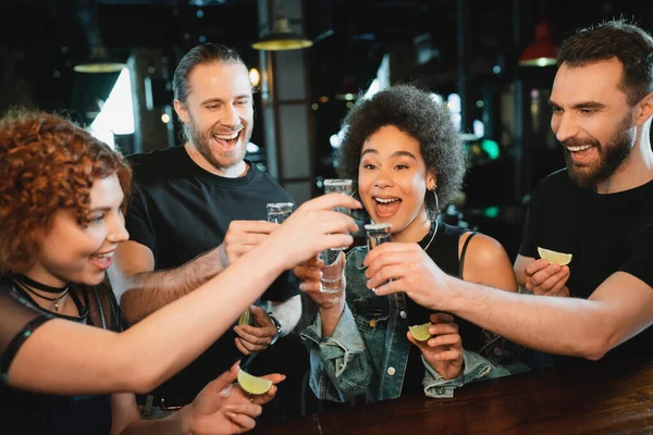 Cheerful interracial friends holding tequila and lime near stand in bar — Stock Photo
