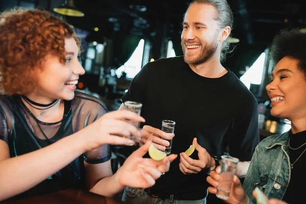 Mujeres multiétnicas positivas sosteniendo tequila y lima cerca del hombre barbudo en el bar - foto de stock