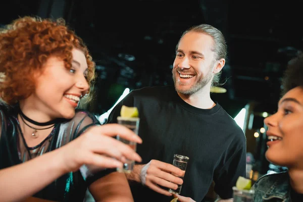 Sonriente hombre barbudo mirando borrosa interracial amigos con tequila en el bar - foto de stock