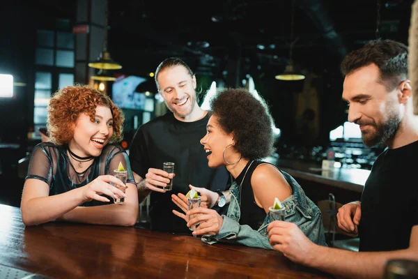 Улыбающиеся межрасовые женщины, держащие стаканы текилы рядом с бородатыми друзьями в баре — стоковое фото