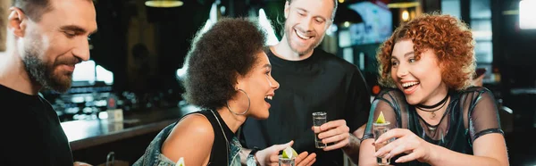 Amigos interracial positivos hablando y sosteniendo tiros de tequila en el bar, pancarta - foto de stock