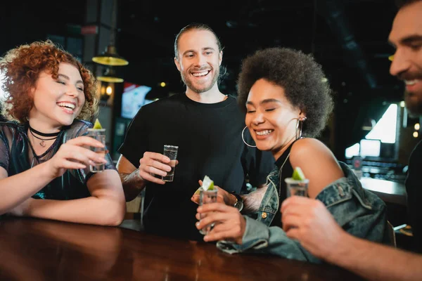 Cheerful multiethnic friends holding tequila shots with lime in bar — Stock Photo